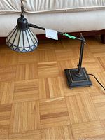 Full view of the desk lamp showing Tiffany style stained glass shade, adjustable arm, and square base on wood floor.