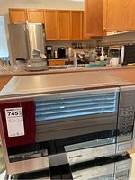 Panasonic microwave oven placed on a kitchen counter with wooden cabinets and stainless steel refrigerator visible in the background, showing front and top sides of the microwave.