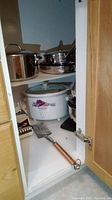 Lower cabinet shelf containing stainless steel pots, small crock pot with floral design, metal spatula with wooden handle, and some smaller items.