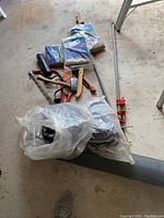 Photo showing assortment of garden and hand tools including wire mesh roll, bird feeder mesh tube, burlap sacks, hand saw, hatchets, tarp, and pruning shears on a concrete floor.