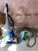 Full view of Shark upright vacuum, Bissell steam cleaner, hose attachment, detergent bottle on floor in basement.
