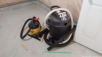 Photo showing black Craftsman wet/dry vacuum and yellow Stinger wet/dry vacuum with hoses on concrete floor.
