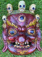 Front view of red lacquered carved Tibetan ceremonial mask with three eyes and decorative skulls on top. Shows the detailed facial features and paint condition.