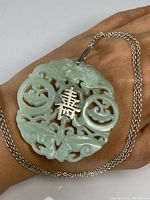 Close-up of the carved jade disc pendant showing detailed Chinese motifs and central character with attached silver necklace on a hand.