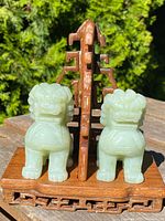 Front view showing pair of pale green carved jade Foo Lions seated next to each other on a hardwood stand with Chinese decorative carving.