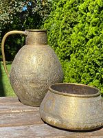 Antique Eastern brass bowl and pitcher displayed outdoors on wooden table, showing intricate chased patterns and overall shape.