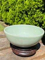 Light green celadon porcelain bowl on wooden stand photographed outdoors with greenery background, showing bowl profile and surface finish.