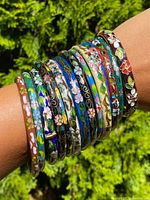 Brass arm adorned with 13 antique cloisonne bracelets featuring a range of colors and floral designs, some with visible clasp elements, shot in bright outdoor lighting.