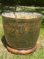 Full front angle of large antique bronze and gilt jardiniere on wooden stand outdoors on grass.