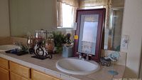 Bathroom sink countertop with the framed print leaning against the mirror, two brown glass candles in decorative holders, a faux plant in a metallic pot, two small dispensers, a bathroom scale, and a blue decorative stand.