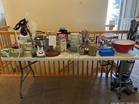 Wide shot of tabletop items including glass cloche, plastic bowls, crockpot, toaster, and various kitchen and barware items arranged on a white folding table.