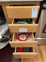 Four wooden kitchen drawers opened showing various kitchen tools, flatware, and linens. Items visible include metal colanders, plastic funnels, red apple corer, and utensils.