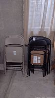 Two stacks of folding chairs, beige metal on the left and black padded on the right, in a basement setting near a window.