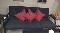 Front view of black metal futon with black cushioned pad and three red throw pillows placed on it.
