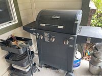 Black Brinkmann outdoor grill with curved lid, three control knobs, and side burner, positioned next to wall.