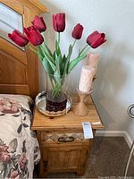 Full view of nightstand next to bed with faux red tulips in glass vase on decorative silver mirror tray and two wooden candleholders with pink candles