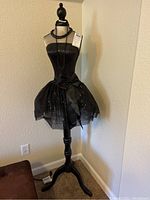 Full view of black mannequin dressed in strapless sequined party dress with tulle skirt and bow, standing on black pedestal.