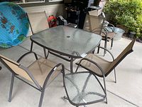 Full patio set showing square glass-top table with umbrella hole, six beige stacking chairs, and square side table with glass top.