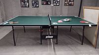 Front view of a green Prince ping pong table with net and some equipment on top including paddles and balls.