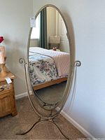 Full view of free-standing oval mirror with metal frame and base standing on carpet in a bedroom setting.