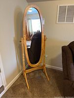 Front view showing full oval mirror with oak colored wood frame, supported by turned side posts and curved base legs on carpeted floor near wall and brown upholstered chair.