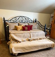 Full view of metal trundle daybed with bedding and pull-out trundle visible underneath, made up with quilted beige bedspread and several pillows in shades of beige, gold, and red.