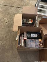 Two cardboard boxes filled with a collection of VHS tapes and music CDs. Visible VHS tapes include standard black plastic cases and some paper sleeves.