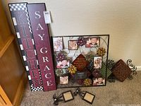 Two tall rectangular wooden plaques with 'KITCHEN' and 'SAY GRACE' text with checkerboard pattern leaning against the wall next to a large square metal photo frame with multiple glass photo slots and metal flowers, plus two small metal photo frames on the floor.