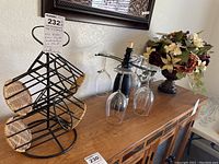 Black metal wine rack with wicker holders, wire stemware display with 4 glasses, and artificial floral arrangement on wooden cabinet top.