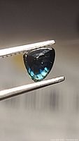Close-up of deep blue trillion cut sapphire held by tweezers showing color and cut details.