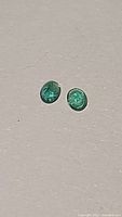 Pair of small oval cut green emerald gemstones on neutral background.