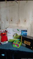 Wide view showing all items in the lot: Elmo toy, dinosaur foam chair box, xylophone toy, and unopened Despicable Box.