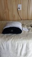 Photo of the foot warmer plugged in and resting on a beige surface against a wall outlet showing power connection.