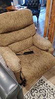 Front view of the brown textured fabric power lift recliner chair showing padded cushioned armrests, backrest, and seat with a remote control on the seat.