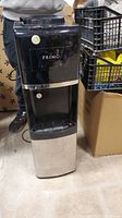 Full view of a tall free-standing black and silver Primo water cooler with taps and drip tray, positioned next to boxes and storage crates.