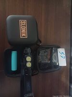 Photo showing the Slonik headlamp alongside a turquoise battery, black carrying case with the Slonik logo, and a mesh pouch with accessories, all arranged on a wooden surface.