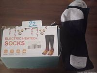Photo showing black and grey electric heated socks lying next to original box with product information and USB charging cable.
