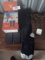 Photo of electric heated socks laid flat on a surface with two black battery packs, a white plug-in charger with cord, and the original branded orange and blue box showing branding and usage graphics.