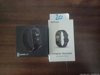 Photo showing two boxed fitness trackers, Kore 2.0 on the left and Befitrack on the right, placed on a wooden surface with a label '20' on the Befitrack box.