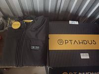 Folded black PTAHDUS heated jacket with zipper and logo, next to original branded box.
