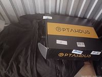 Photo of black heated jacket folded next to black and yellow PTAHDUS branded box with visible size M-5 tag on box.
