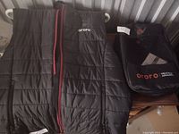 Black Ororo heated vest laid flat with front zipper and pockets visible, next to its carrying bag displayed on a wooden surface.