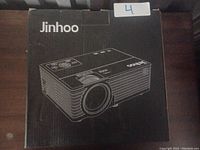 Photo of black cardboard box with Jinhoo branding and projector outline drawing, marked with sticker '4'.
