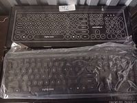 Photo shows Inphic wireless keyboard inside clear plastic sleeve with full key layout and round keys. Its packaging box is behind it with Inphic branding.