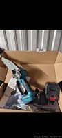 Mini cordless chain saw with a black and blue body, ergonomic handle, and small saw blade; accompanied by a black detachable battery and charger with red latch, inside a cardboard box.