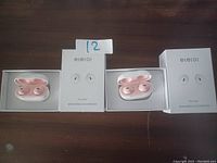 Two sets of mini wireless earbuds in pink charging cases, displayed beside their white original boxes on a dark surface.