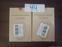 Two boxed rechargeable hand warmers shown side by side on a wooden surface with a sign indicating lot number 44.
