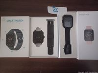Photo showing two smart watches in their original boxes along with an extra black band strap. One watch has a black band and the other has a pink band in box.