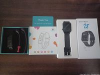 Smart watch and accessories neatly arranged: watch in the box insert, charging cable with red tip, instruction manual, thank you card, and product box with the image of the smart watch.