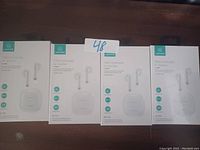 Four boxed USAMS TWS earbuds arranged side by side on a dark surface, each box shows image of white Bluetooth earbuds and charging case with product features.